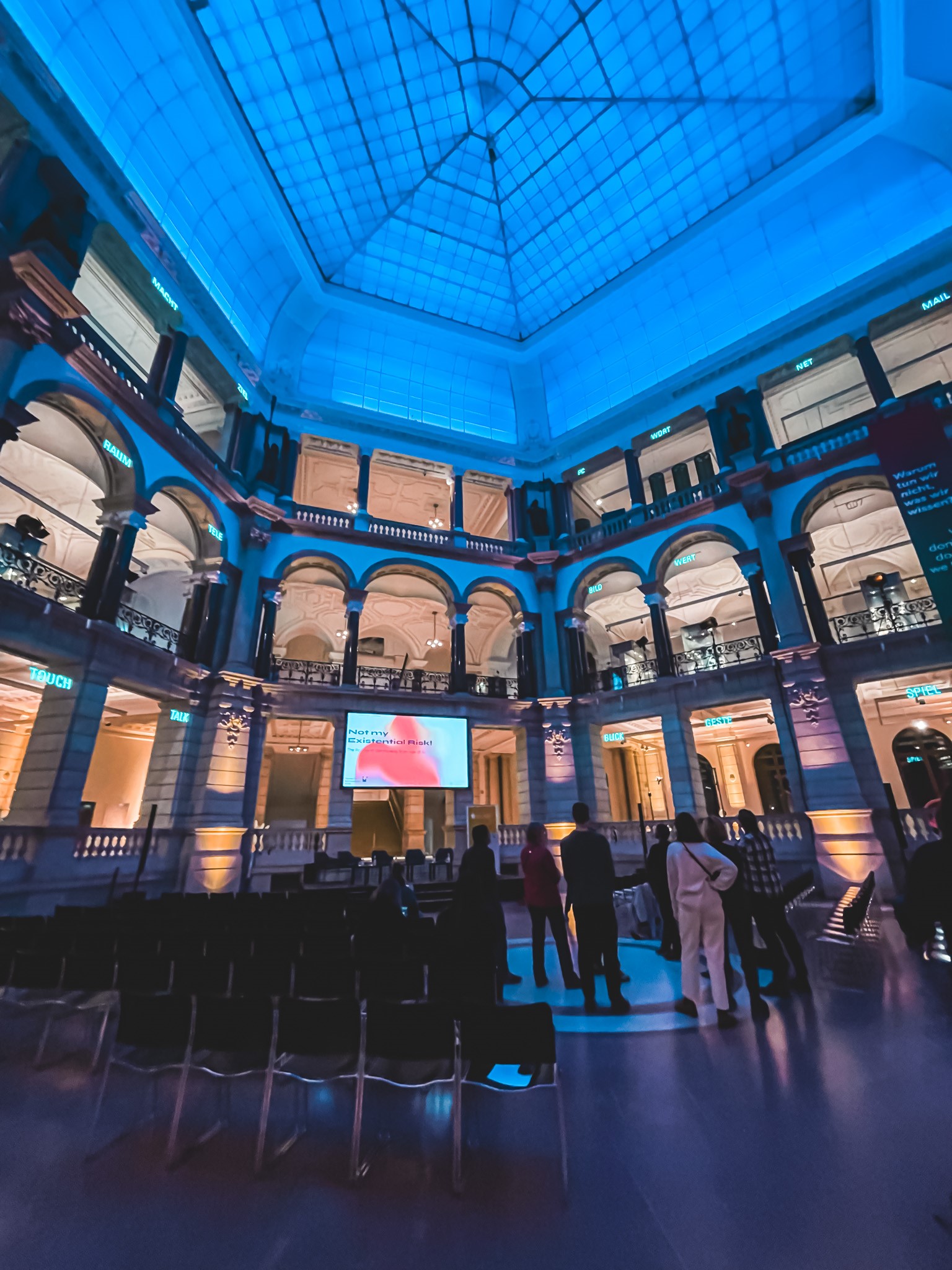 Photo depicting indoor of a building with round arched walls and a glass roof atop of a hexagonal space. People in a hall are watching the screen and socialising.