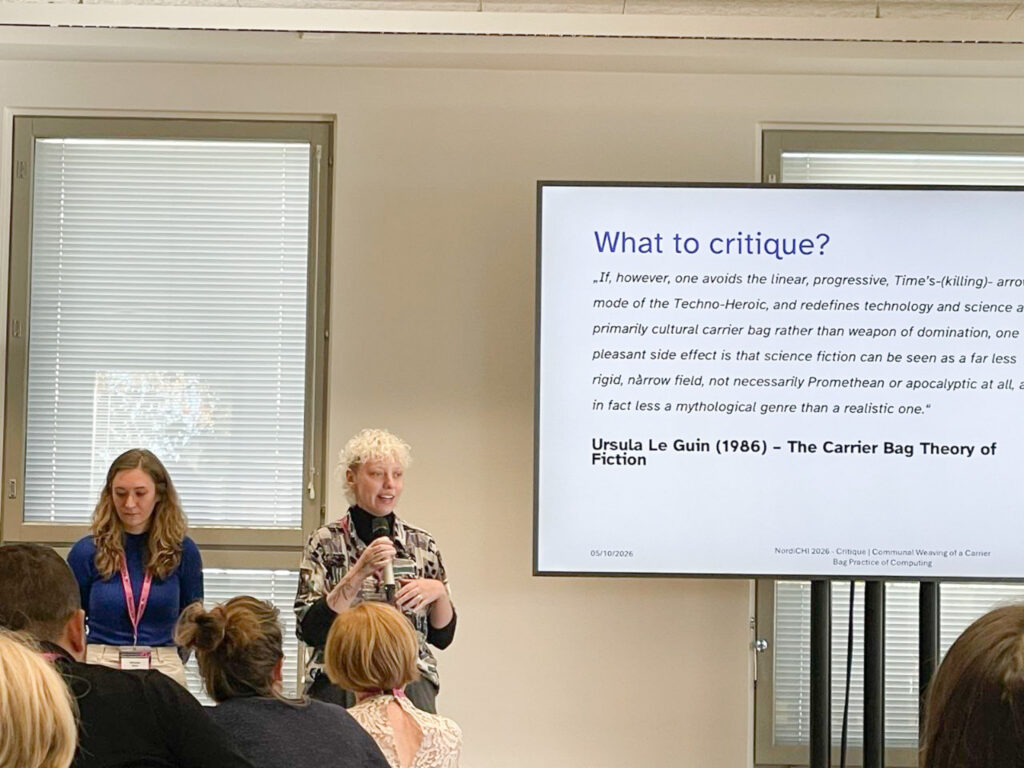 two people presenting at the front of a university classroom, a screen is showing a powerpoint presentation, the slide title reads "What to critique?"