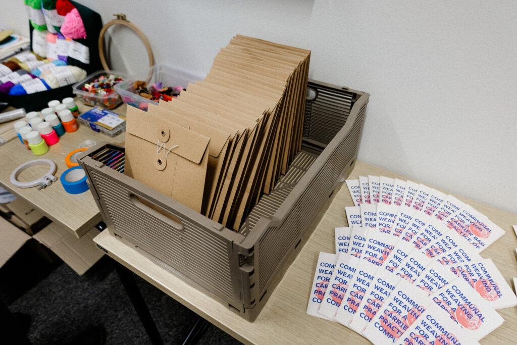 brown cardboard envelopes in a box on a table, next to the envelopes are a selection of zines with the words "COMMUNAL WEAVING FOR A CARRIER BAG PRACTICE OF COMPUTING" on it.