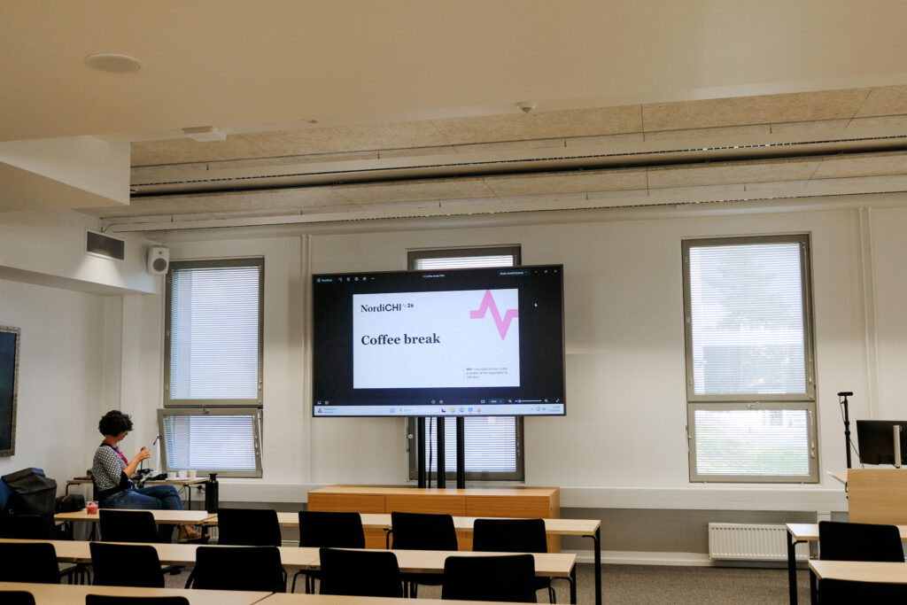 Photograph of a university class room, a screen displace a powerpoint slide with the "NordiCHI26" logo and the words "Coffee break"; in the corner of the room is a person sitting and knitting