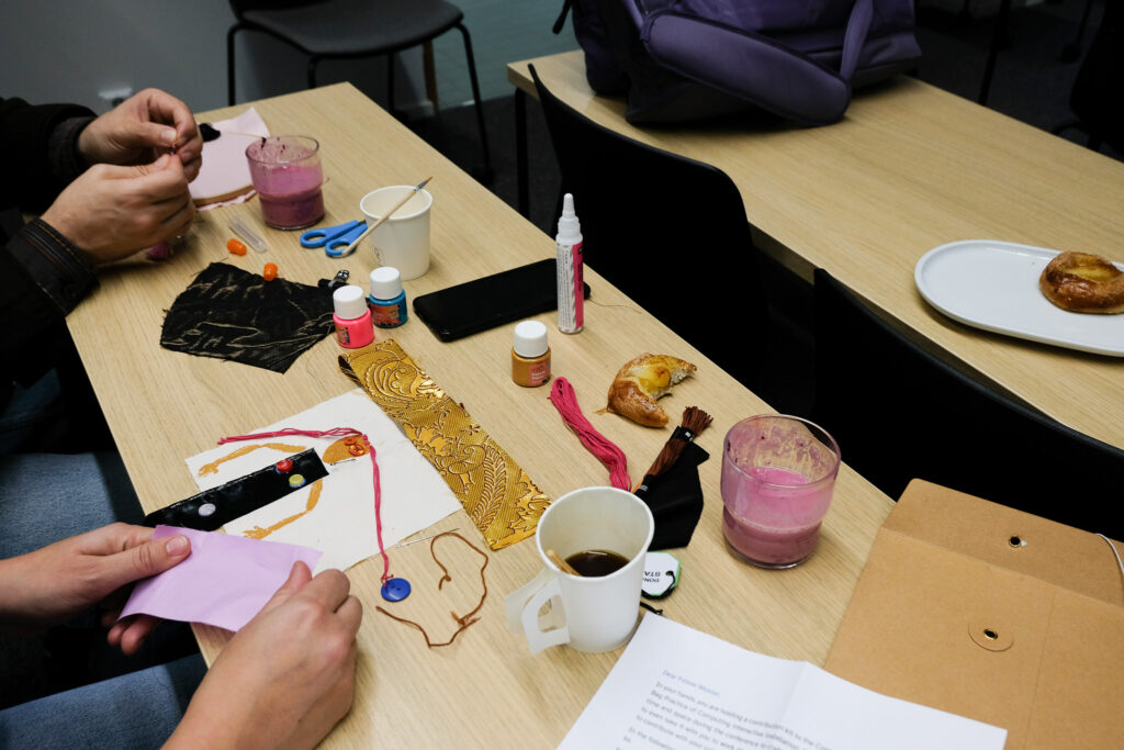 A view of two people's hands crafting at a table; on the table there are coffee cups, fabric, paint, string, and glue