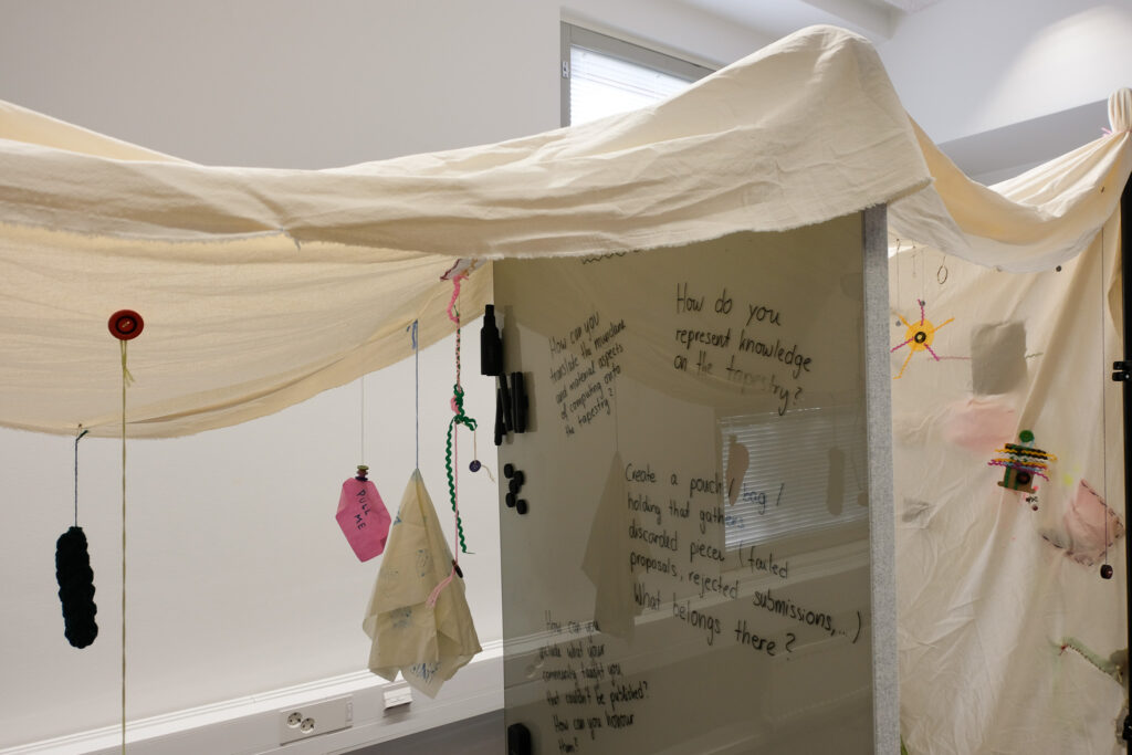 A piece of cloth hung over a whiteboard wall, there are more pieces of fabric attached, hanging from it